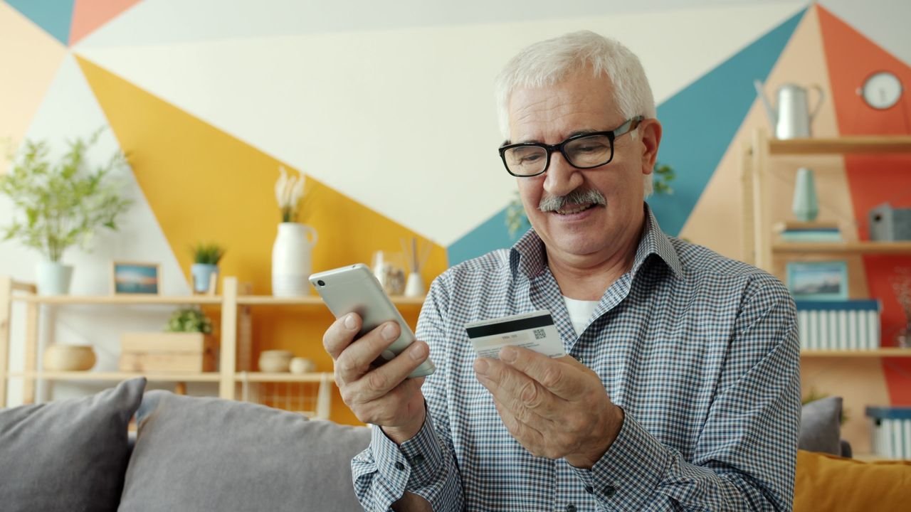 heros-img Senior man using smartphone and credit card for online shopping indoors.